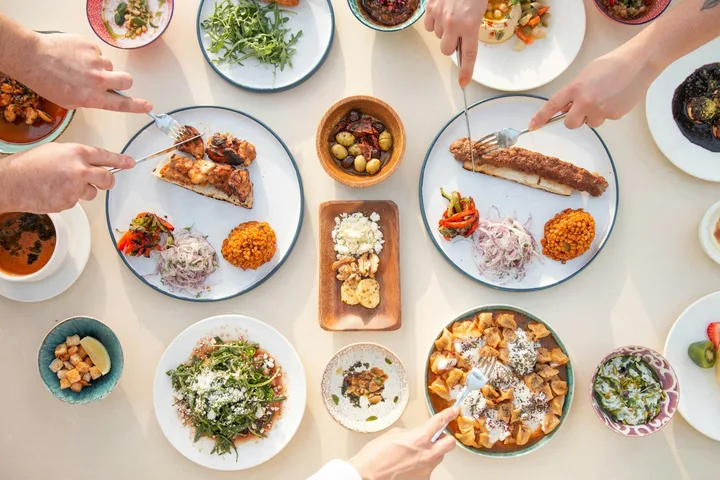 Overhead view of many shared dishes on a table, with diners reaching in with forks