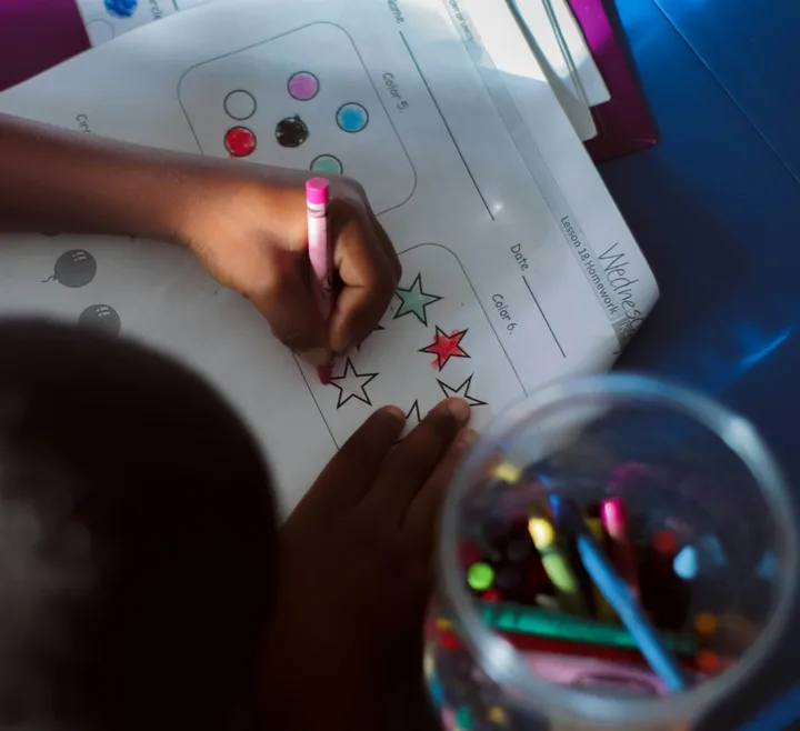 A child colors in stars on a worksheet with a pink crayon, next to a glass jar of crayons