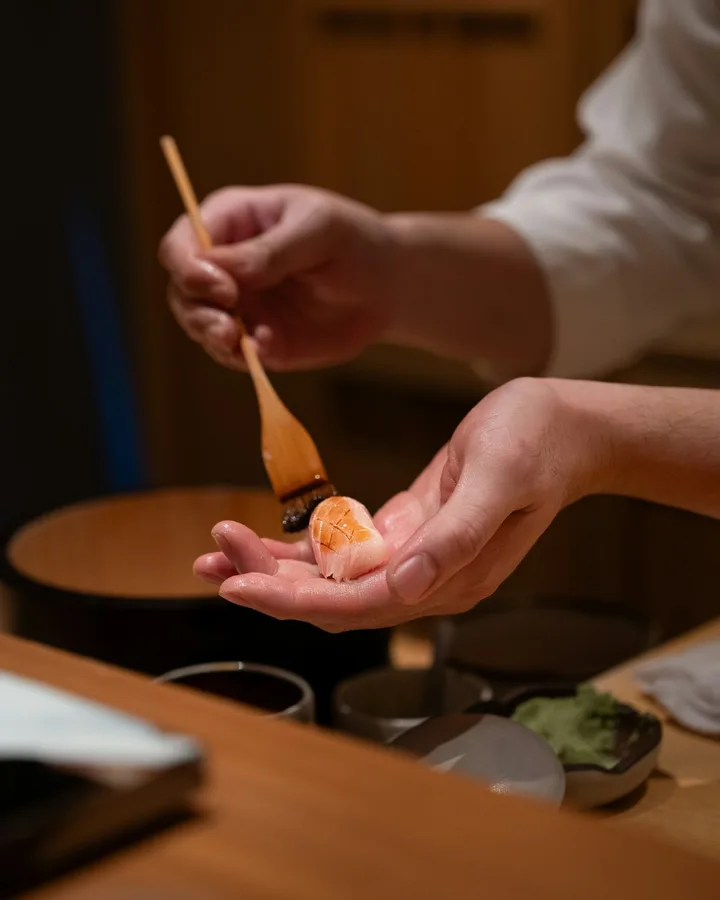 Sushi chef brushing sauce onto a piece of nigiri at the counter