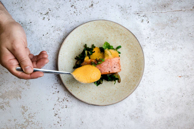A chef finishing a salmon dish with sauce and fresh greens
